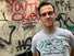 A man wearing a gray t-shirt with a green graphic and glasses, standing in front of a concrete wall covered in graffiti.