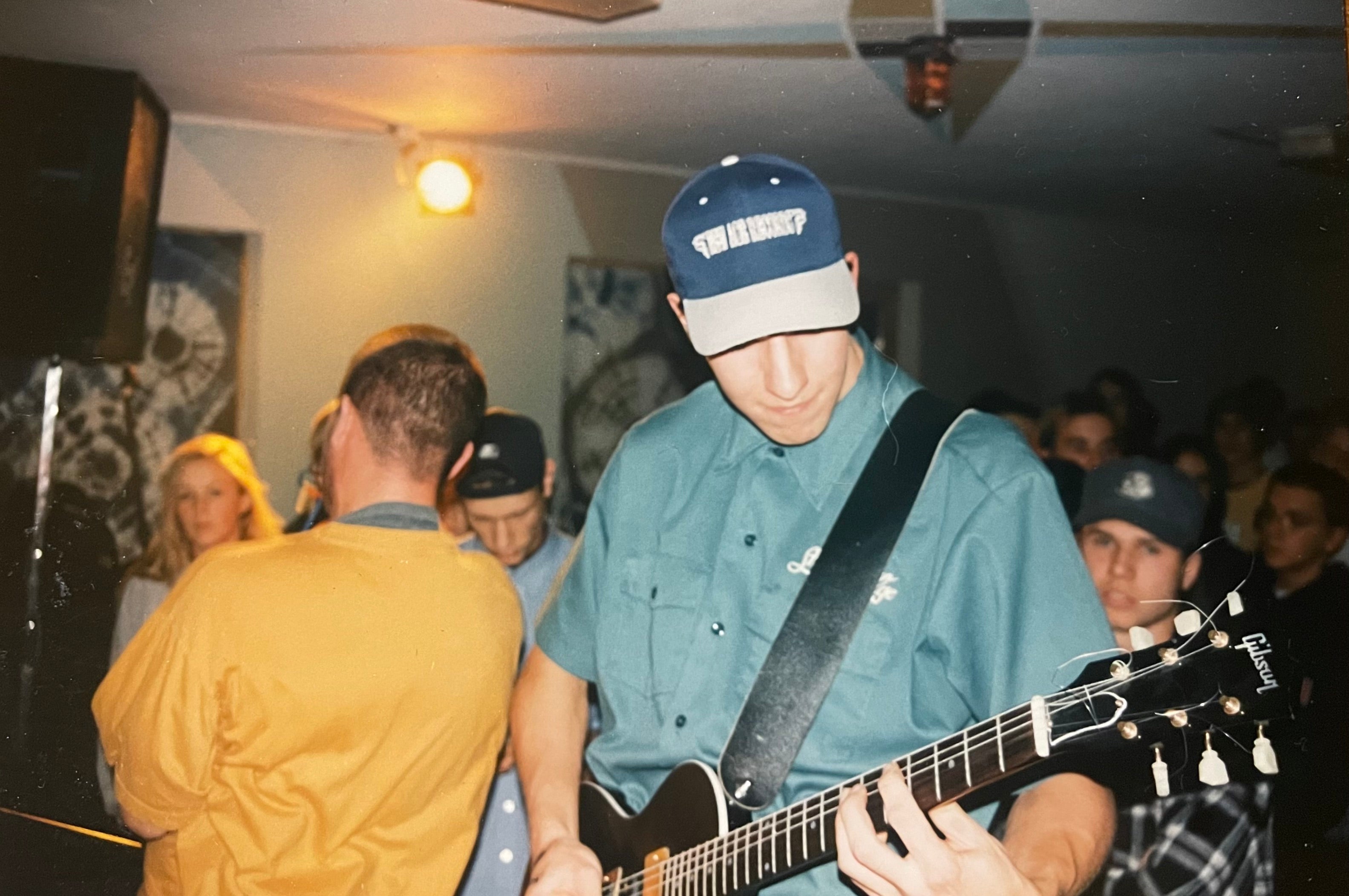 A person wearing a blue and white baseball cap with 'NEW AGE RECORDS' printed on it, a teal button-up shirt, and a black guitar strap, playing a black electric guitar in a crowded indoor venue.