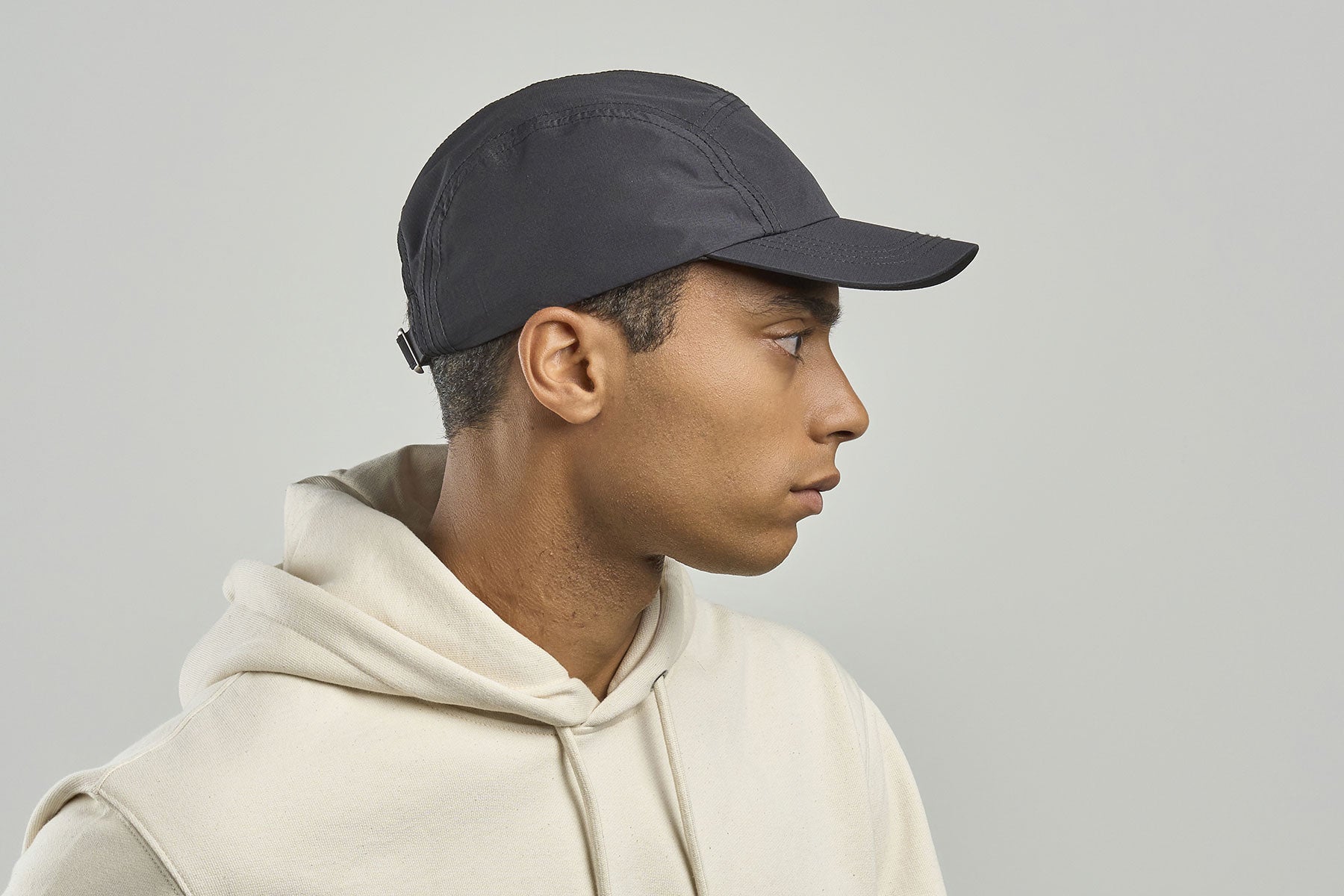 A man in profile wearing a dark charcoal gray 5-panel cap and a cream-colored hoodie, set against a solid light gray background.