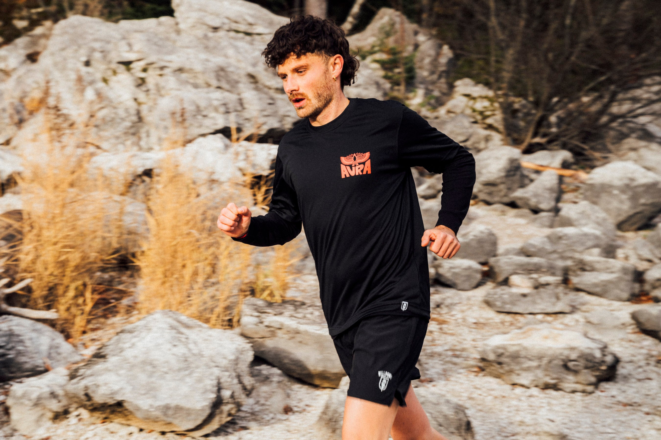 Man running outdoors on rocky terrain wearing a black long-sleeve shirt with red text and black shorts.