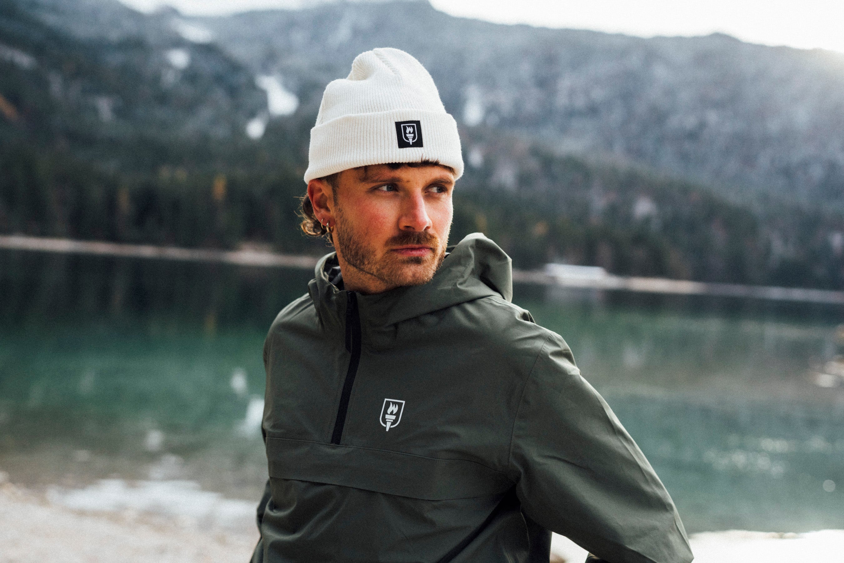 Man wearing a gray jacket and white beanie with a logo, standing by a lake with mountains in the background.