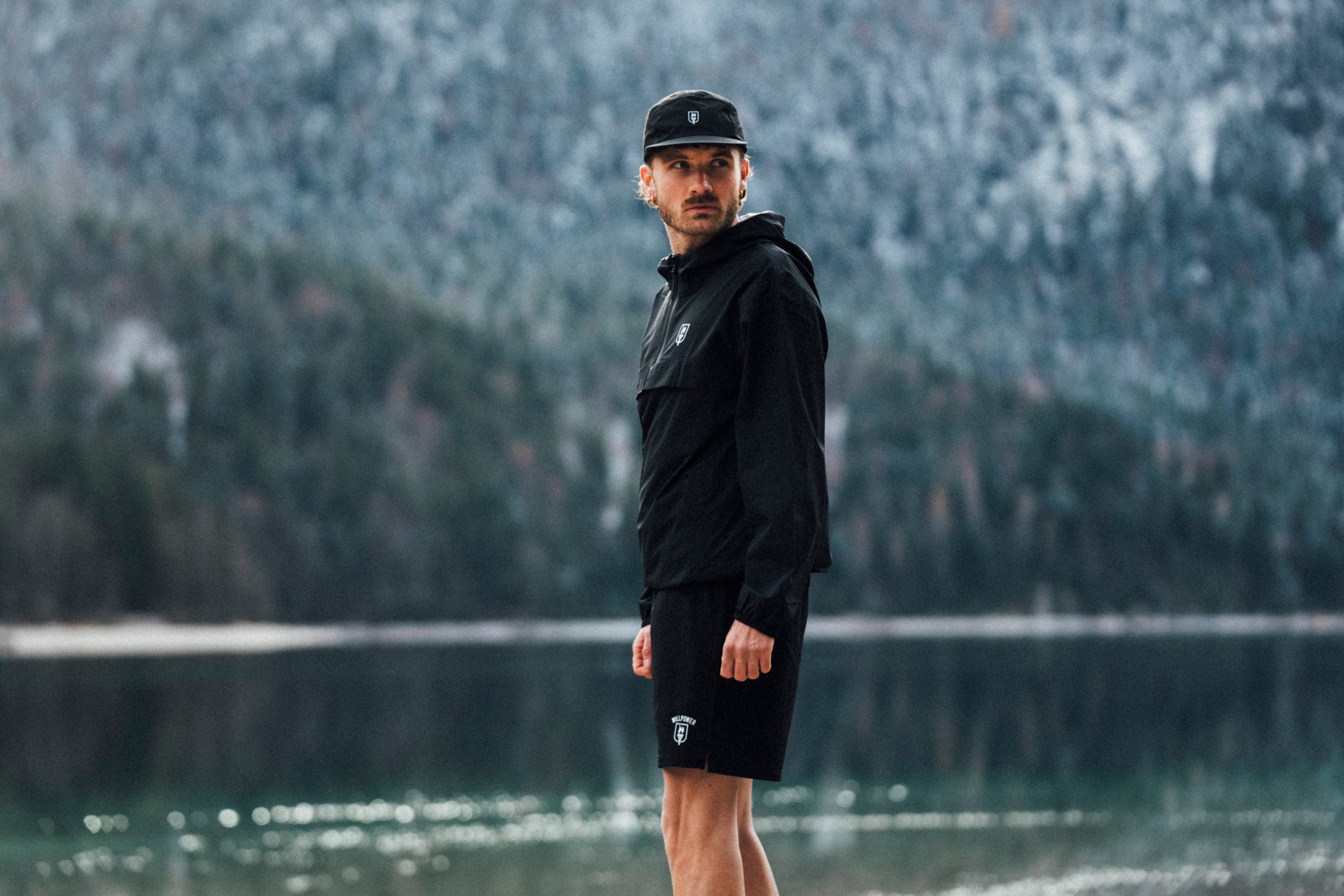 Man in black jacket and shorts standing by a lake with a forested background