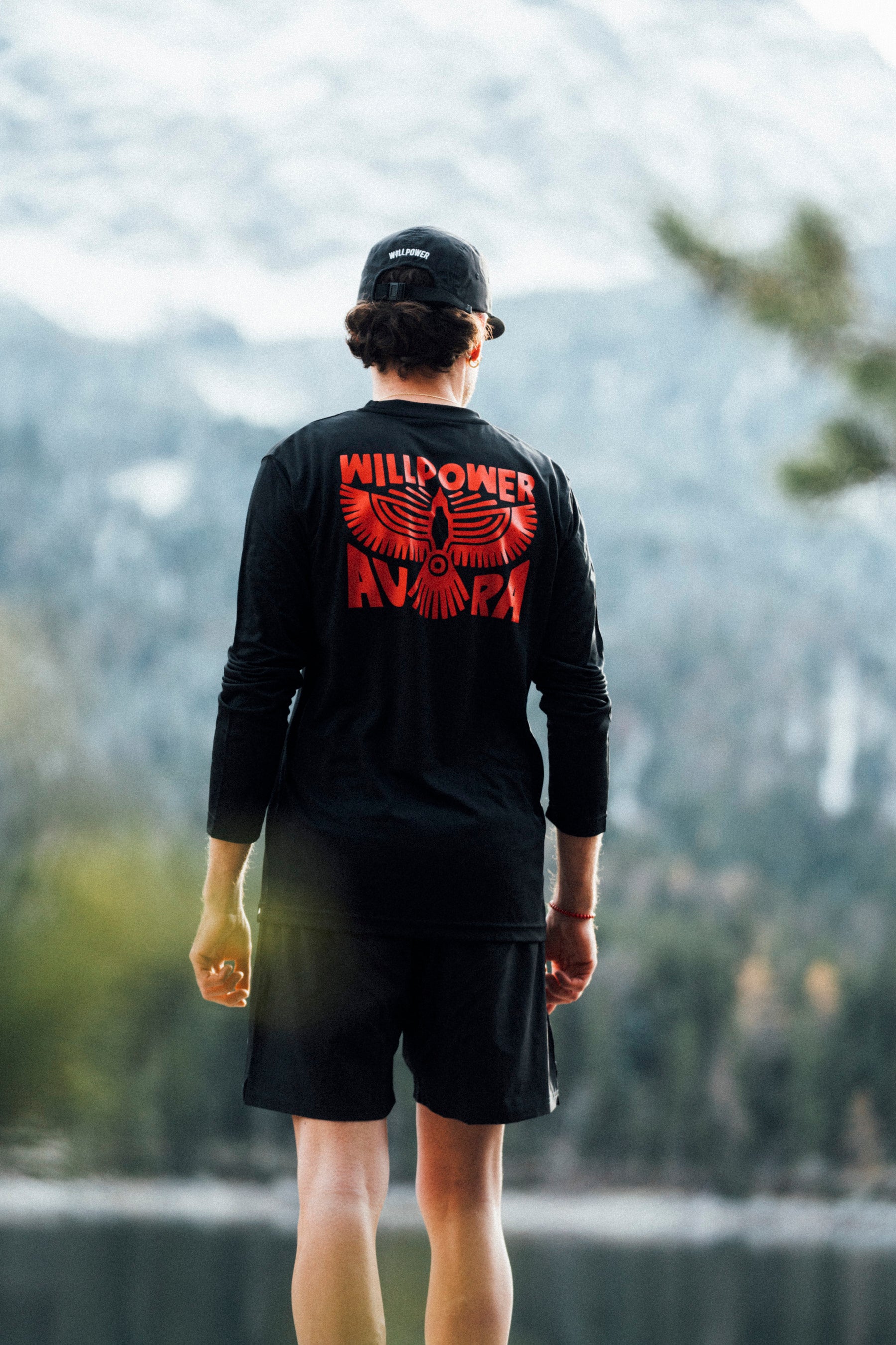 Person wearing a black long-sleeve shirt with red text and graphics, standing in a natural setting with trees and mountains.