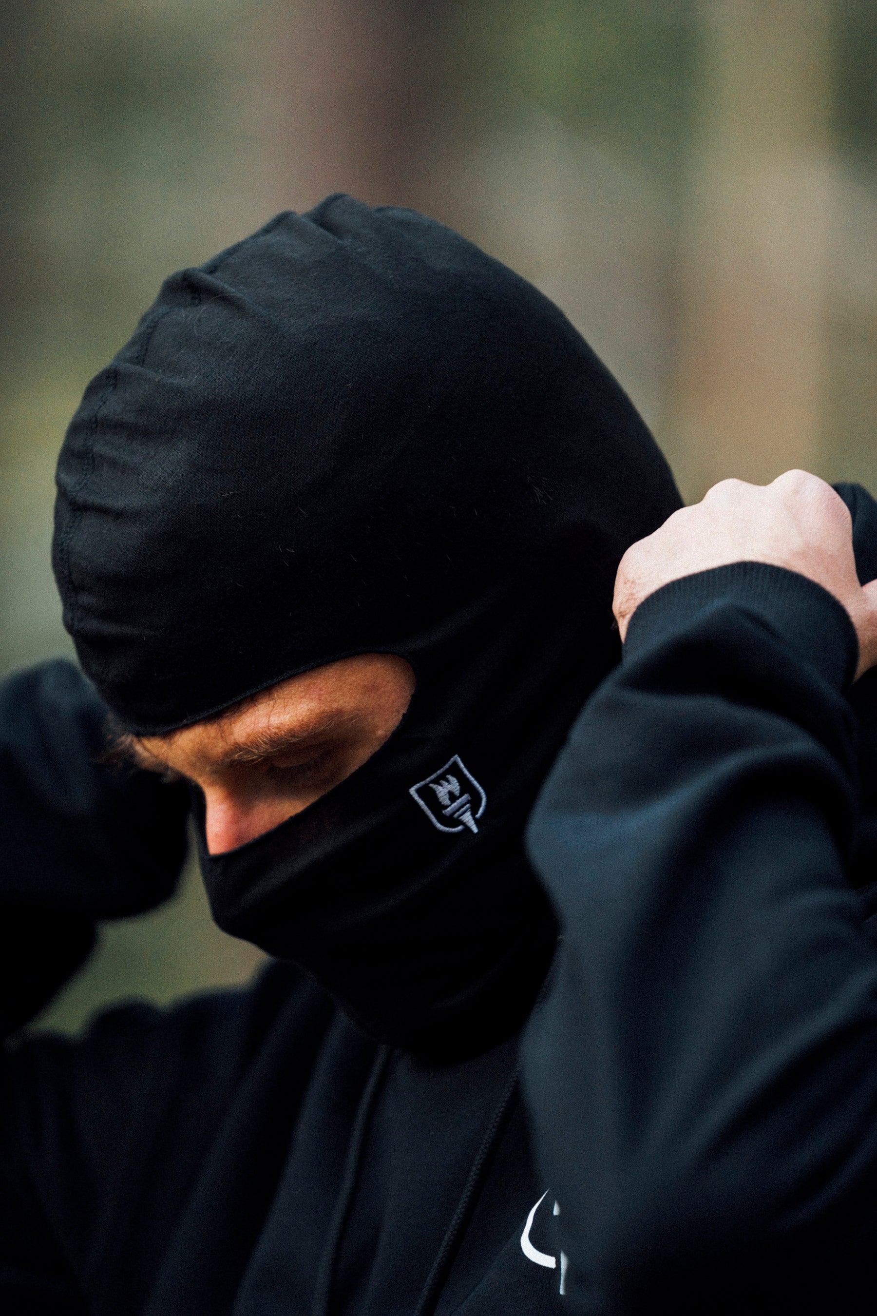 Person wearing a black balaclava with a logo, blurred background