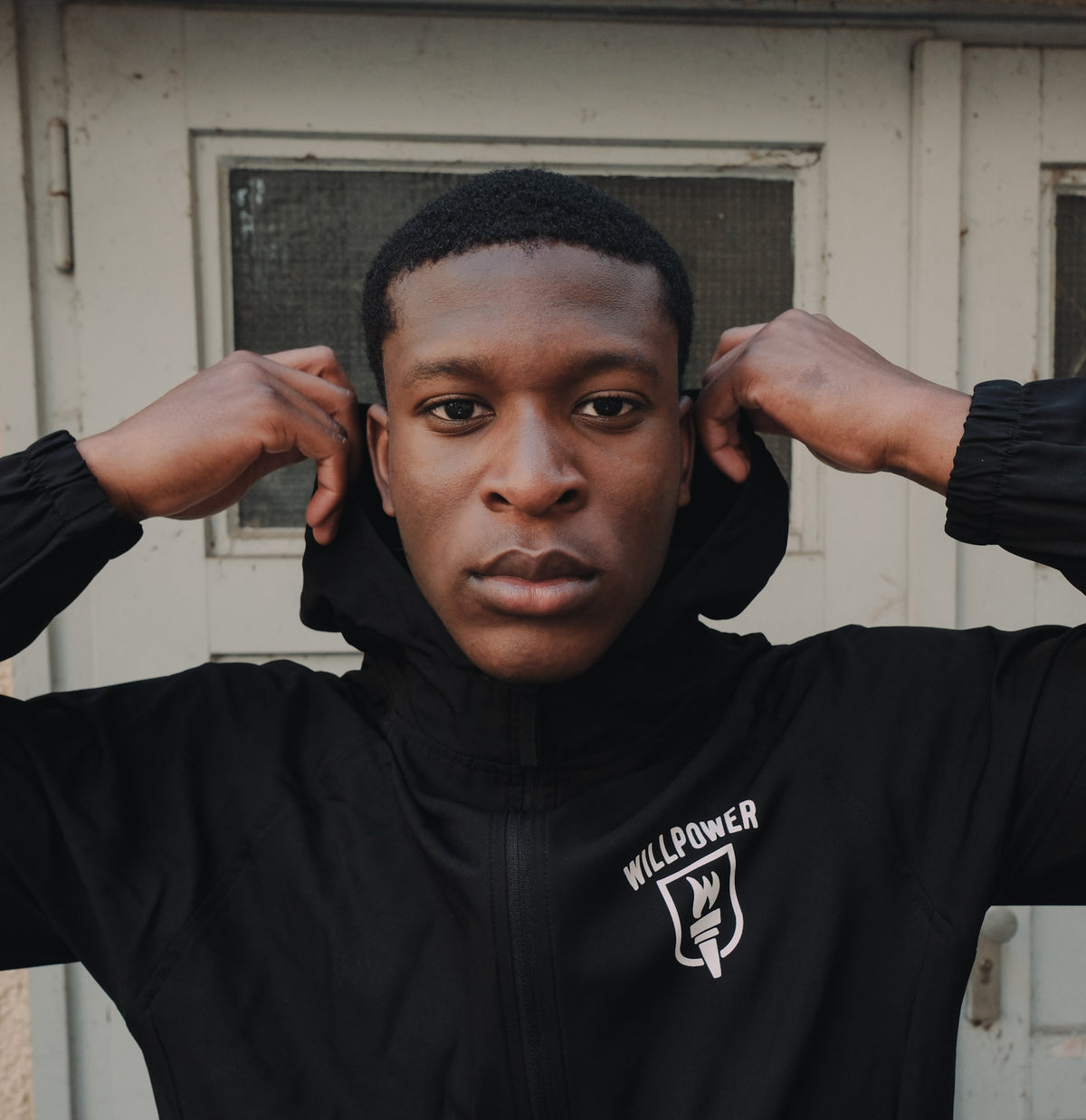 A man wearing a black zip-up hooded jacket with a white 'WILLPOWER' shield logo on the chest, posing with his hands behind his ears in front of a weathered white door.