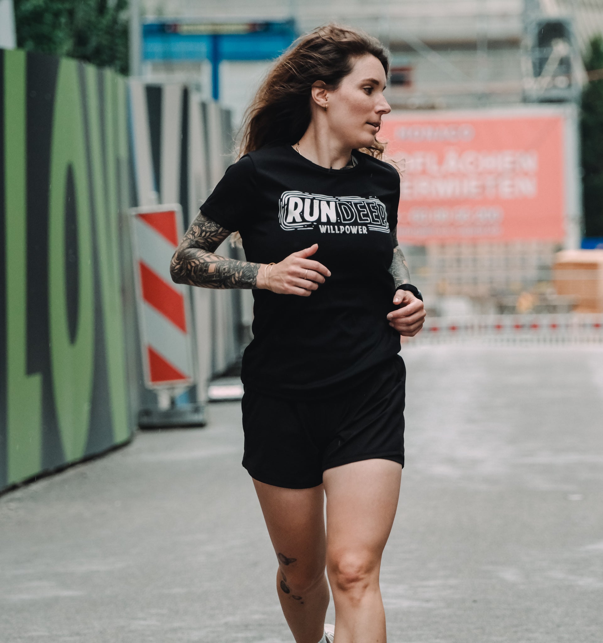 A woman running outdoors wearing a black short-sleeve t-shirt with 'RUN DEEB WILLPOWER' printed in white across the chest, paired with black shorts.
