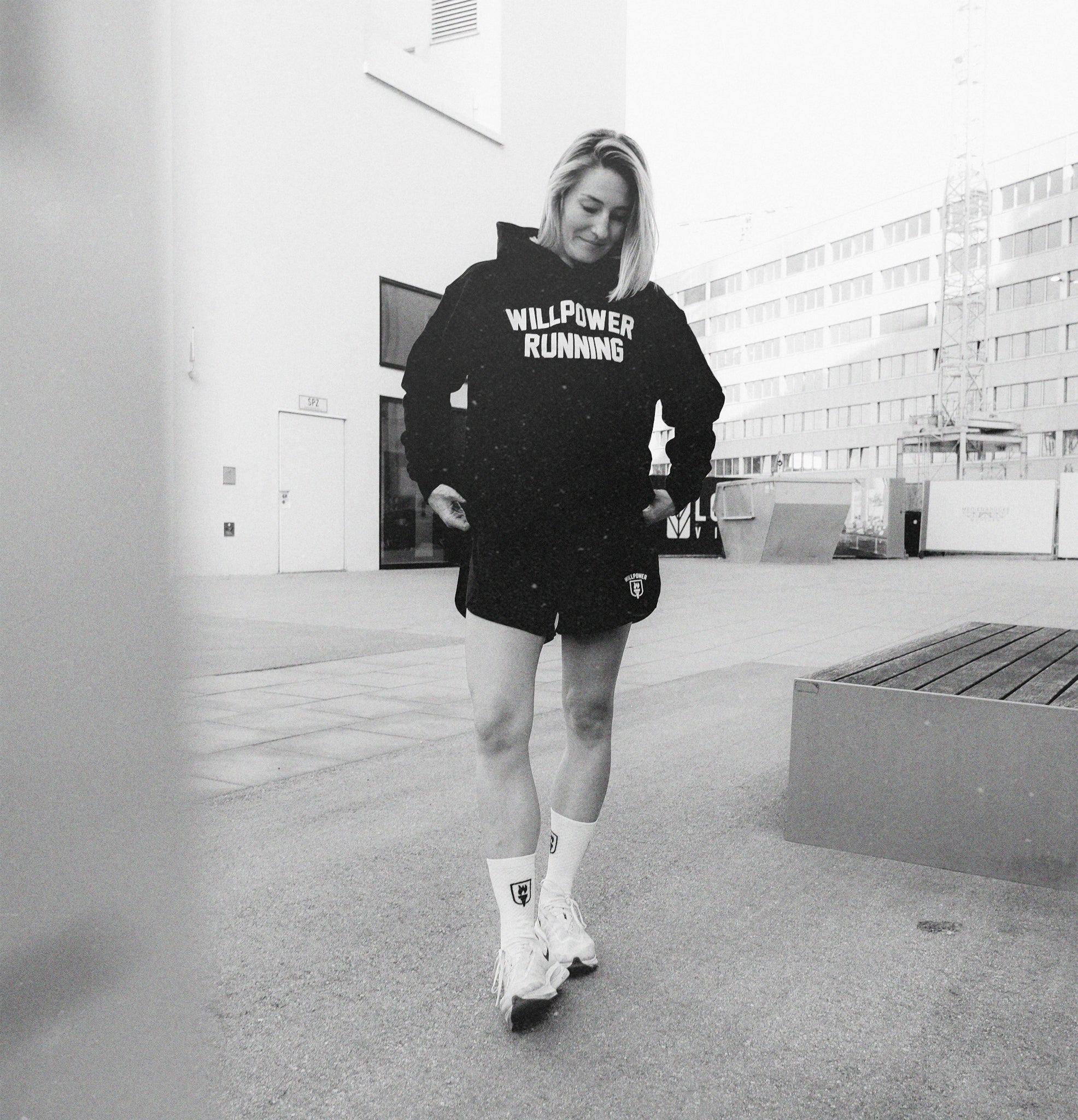 A woman wearing a black hoodie with 'WILLPOWER RUNNING' printed in white on the chest, black athletic shorts, and roller skates, captured in a black and white outdoor urban setting.