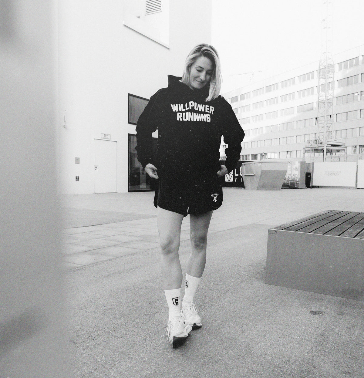A woman wearing a black hoodie with 'WILLPOWER RUNNING' printed in white on the chest, black athletic shorts, and roller skates, captured in a black and white outdoor urban setting.