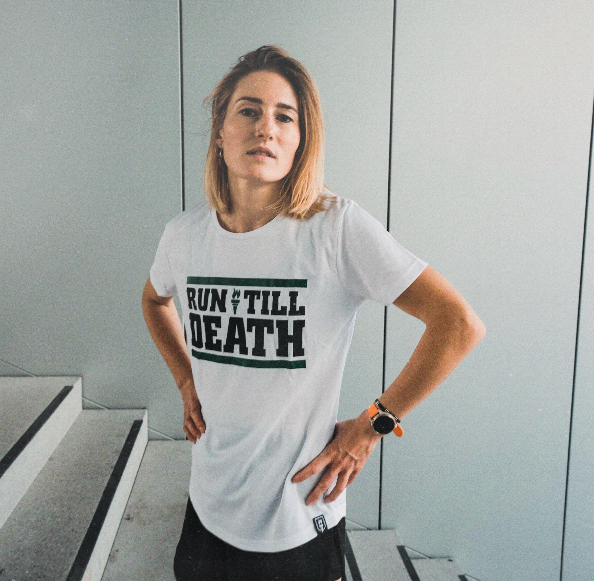 A woman wearing a white graphic t-shirt with the text 'RUN TILL DEATH' printed in black and green, standing in front of a light gray paneled wall.