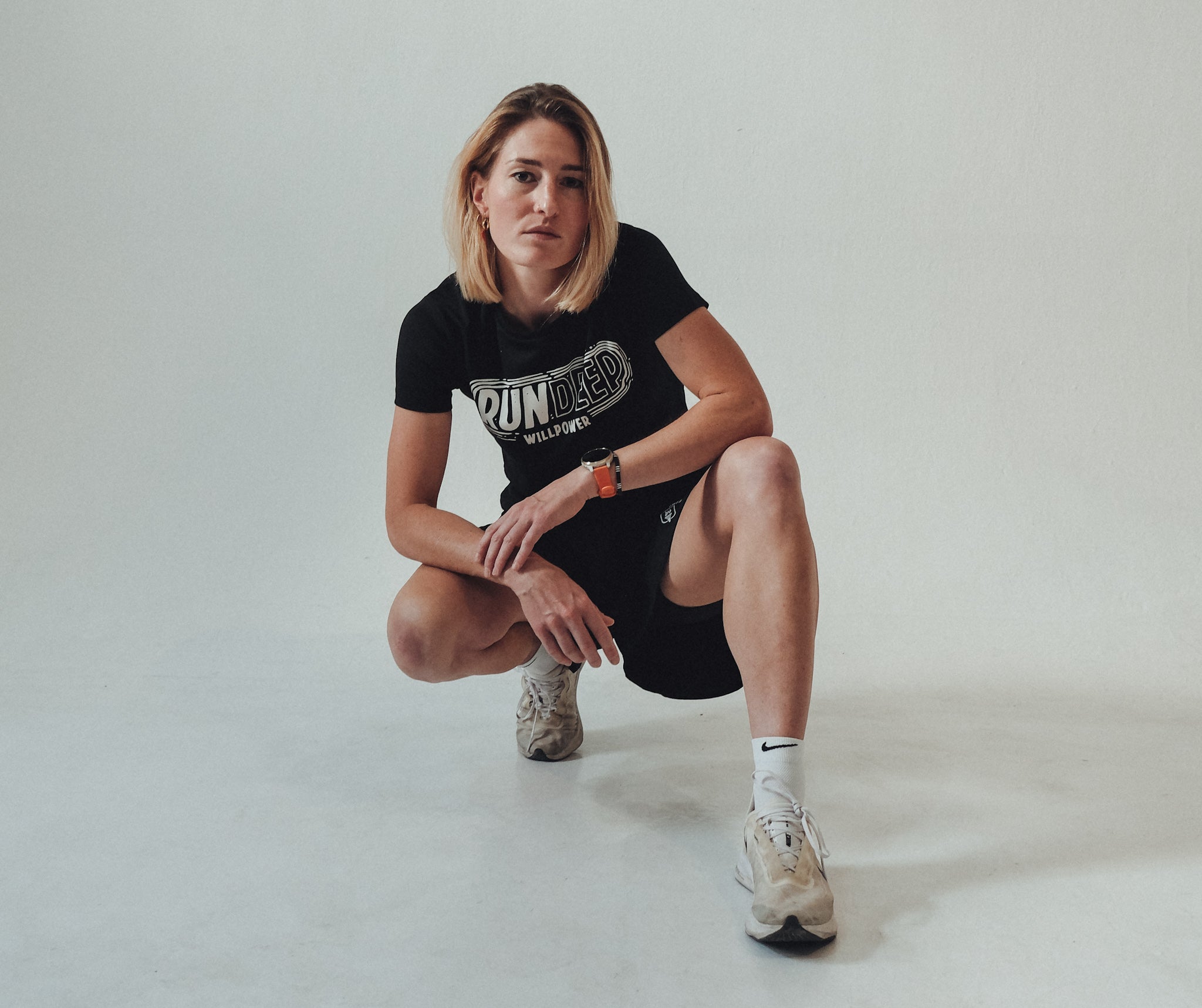 A woman crouching in a studio setting, wearing a black graphic t-shirt with 'RUN DUMP' printed on the front, black athletic shorts, white socks, and beige sneakers.