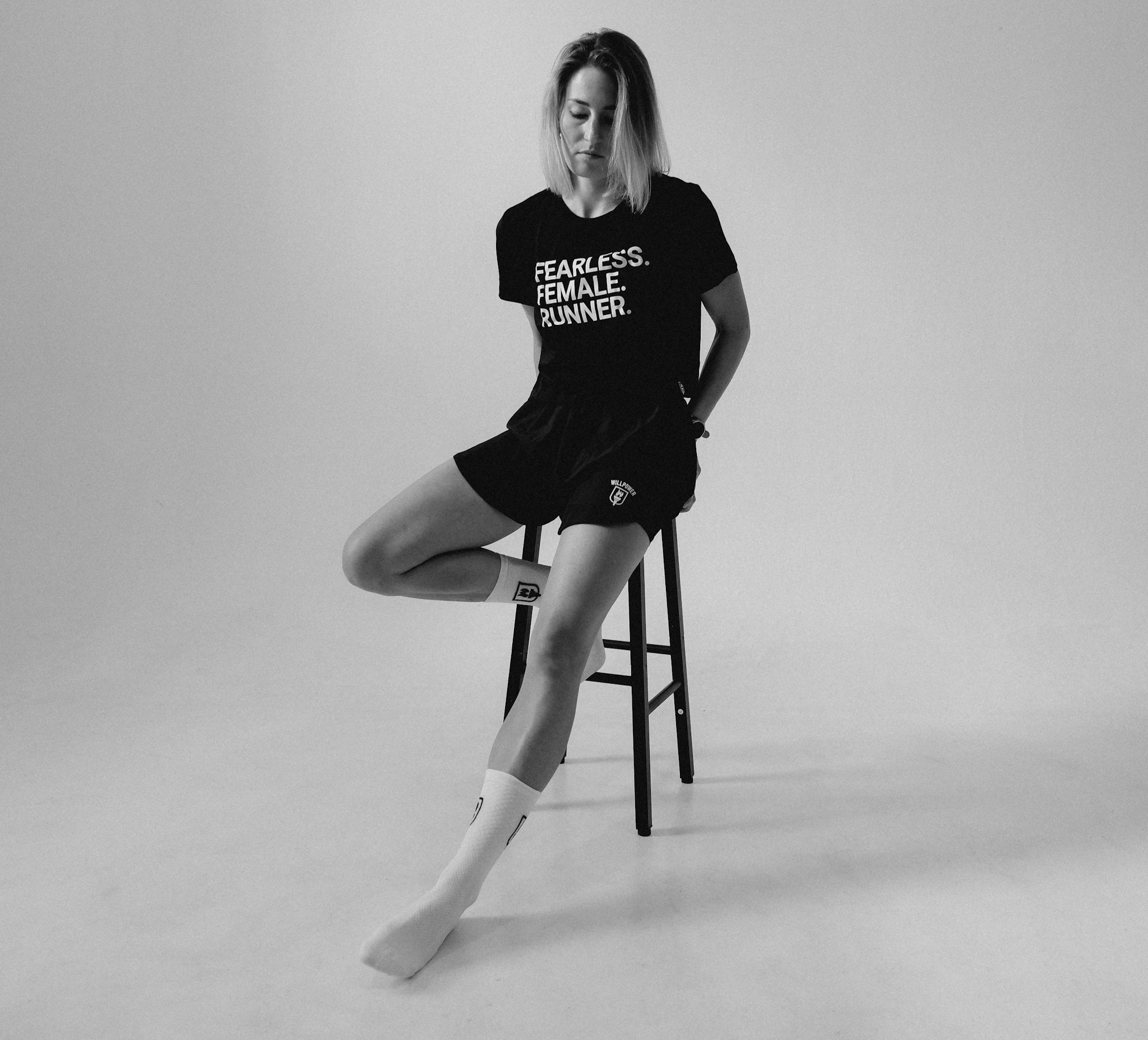 A woman sits on a stool in a studio setting, wearing a black graphic T-shirt with the text 'FEARLESS. FEMALE RUNNER.