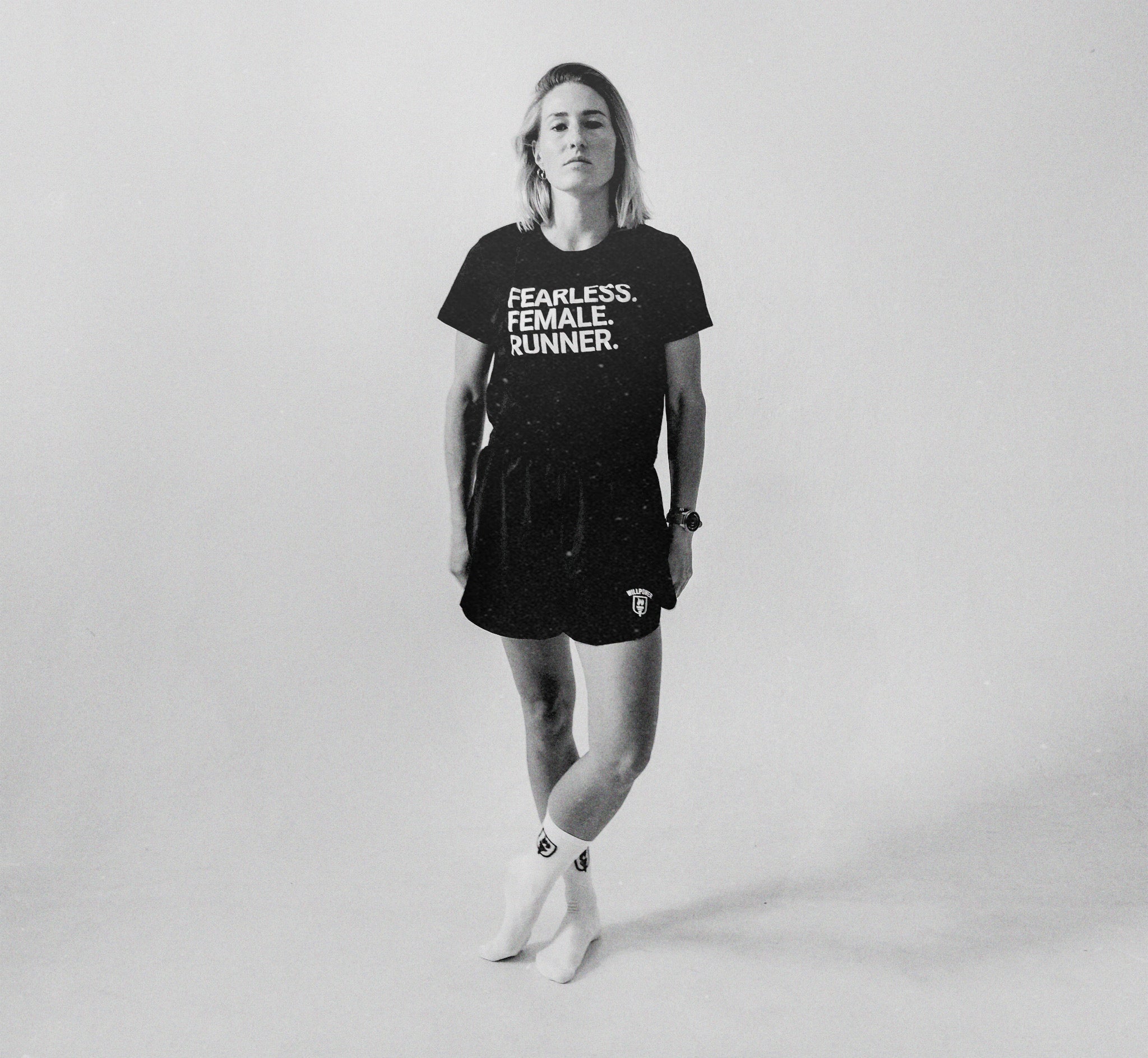 A woman stands in a studio wearing a black graphic T-shirt and matching black shorts with white socks. The shirt has the text 'FEARLESS.
